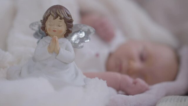 Close-up toy praying angel and blurred carefree infant girl sleeping at background. Relaxed Caucasian baby child resting at home in comfortable bedroom indoors