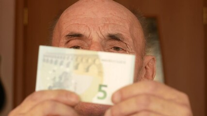 Close-up of a Caucasian pensioner 70 years old holds a 5 euro banknote in front of his face. The elderly man saves, keeps 5 euros. European currency paper money. Selective focus
