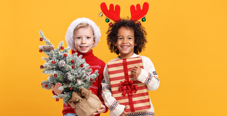 Happy multiracial children wearing santa hat and deer antlers holding snowy Xmas tree and gift box
