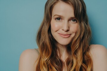 Studio shot of charming shy young woman with wavy brown hair looking at camera with slight smile