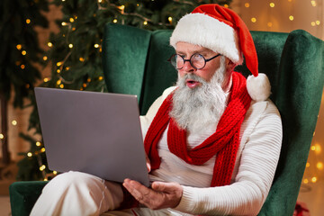 Serious Santa Claus sitting at his home and reading children's email letters , wish list on laptop...