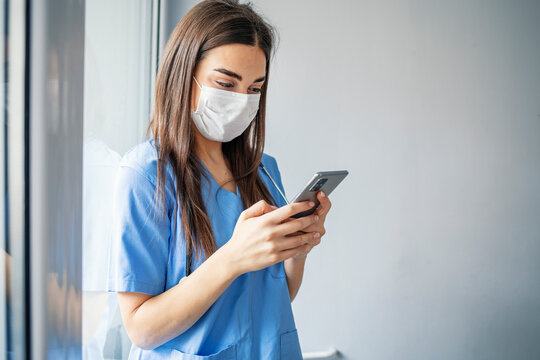 Healthcare Professional Uses Smartphone Phone During COVID-19. A Female Healthcare Professional Wears A Mask While On Break From Her Shift. She Is Checking For Messages On Her Smartphone.