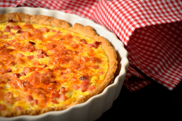 Side view of pie with onion, eggs and bacon. French traditional quiche Lorraine. Baked Custard dough. Pie on white plate with red kitchen towel on black background. Close up of dish
