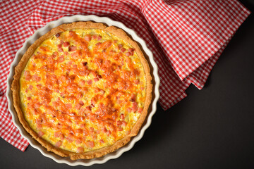 Top view of pie with onion, eggs and bacon. French traditional quiche Lorraine. Baked Custard dough. Pie on white plate with white and red kitchen towel on black background. Directly above shot
