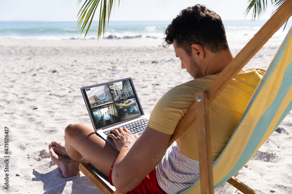 Poster caucasian man on holidays using laptop with views of offices from security cameras on screen
