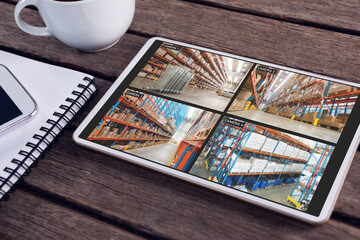 Tablet with views of warehouse from security cameras on screen lying on wooden table