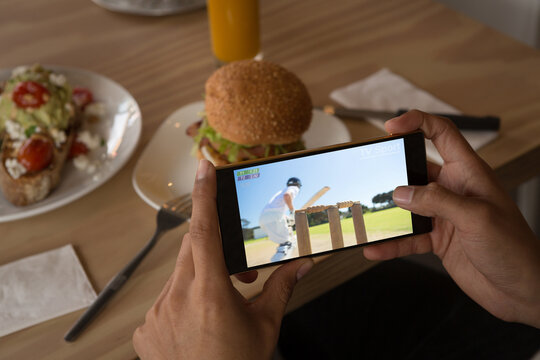 Hands of african american man at restaurant watching cricket match on smartphone