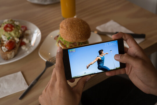 Hands of african american man at restaurant watching male athlete throwing discus on smartphone