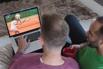 Two diverse male friends at home watching tennis match on laptop - Powered by Adobe