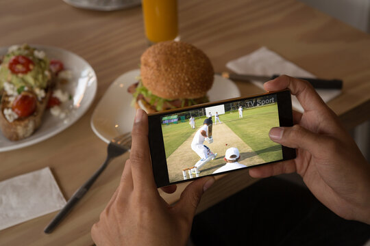 Hands of caucasian man at restaurant watching cricket match on smartphone