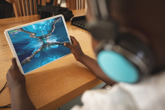 African american boy wearing headphones at home watching swimming competition on tablet