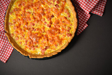 Top view of whole traditional French pie quiche Lorraine with ham, onion and eggs. Backed custard dough served on plate on kitchen towel. Classic recipe. Black background.
