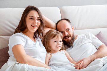 Fototapeta na wymiar Young family play spend time together hugs on couch in morning. Mother Father and Daughter child in white t-shirts pajamas have fun in Morning Routine at home in bedroom. 