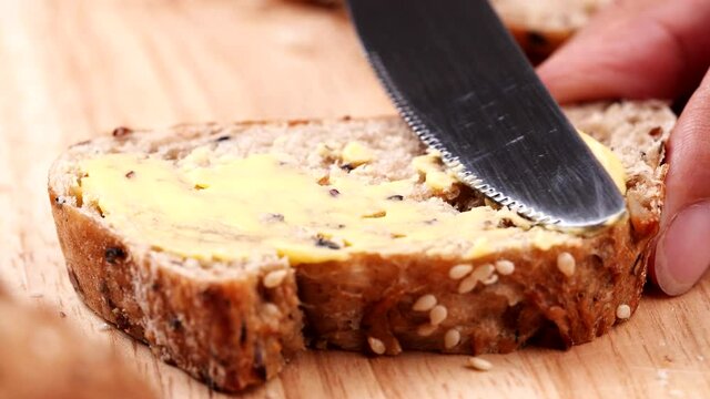 Women holding sliced multigrain bread on wooden table and using butter knife spreading tasty butter cream on fresh bread with seed.