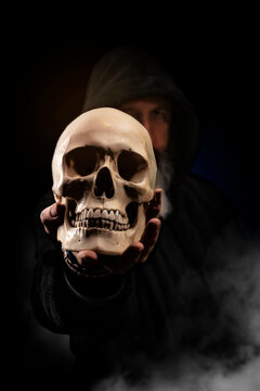 Hooded man holds in his hand a human skull, dark background with smoke. Selective focus.