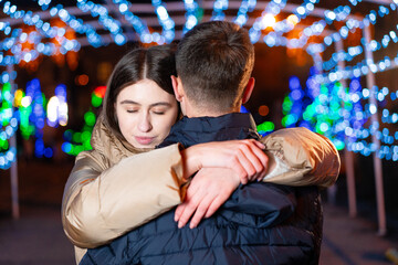 The guy and the girl hug in the winter against the backdrop of the night city, which brightly shines festoon. Girl with closed eyes