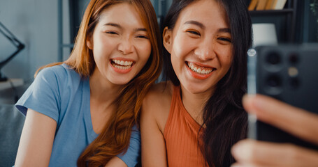Fototapeta premium Teenager Asian women feeling happy smiling relax use smartphone video call in living room at home. Cheerful Roommate ladies video conference with friend and family, Lifestyle woman at home concept.