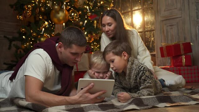 Happy Young Family Using A Tablet Computer At Home Sitting By The Christmas Tree