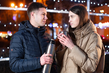 Couple looking at each other and smiling. They drink tea on the street in winter.