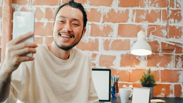 Young Asia businessman using phone talk to colleagues about plan in video call meeting while work from home at living room. Self-isolation, social distancing, quarantine for corona virus prevention. - Powered by Adobe