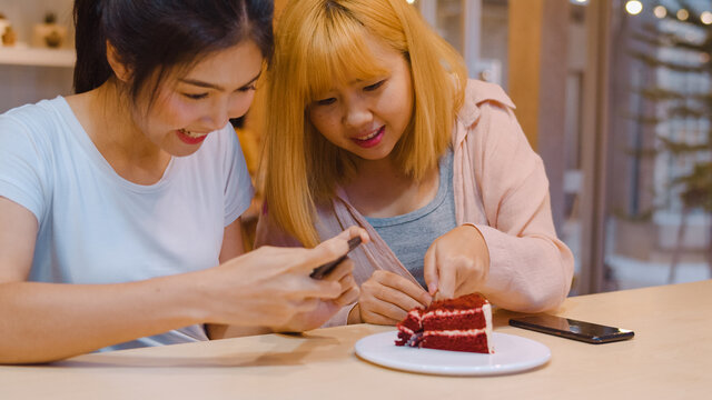 Cheerful young Asia friend using phone taking a photograph food and cake at coffee shop. Two joyful attractive Asian lady together at restaurant or cafe. Holiday activity, or modern lifestyle concept.