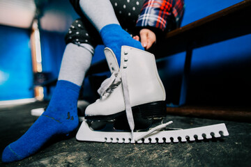 Midsection of caucasian kid in blue socks sit on bench, put on ice skates in changing room in city ice rink. Closed up horizontal shot with focus on skate. Children sport and winter activities concept