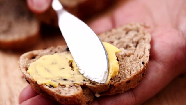 Women holding sliced multigrain bread on wooden table and using butter knife spreading tasty butter cream on fresh bread with seed.