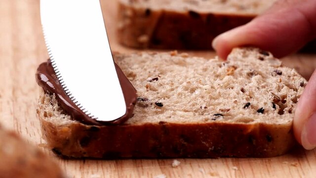 Women holding sliced multigrain bread on wooden table and using butter knife spreading tasty chocolate cream on fresh bread with seed.