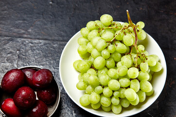 Branch of ripe green grape on plate with water drops and plums in bowl. Juicy fruits on wooden background, closeup. Healthy food on dark kitchen table
