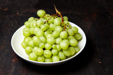 Branch of ripe green grape on plate with water drops. Juicy grapes on wooden background, closeup. Grapes on dark kitchen table with copy space