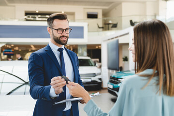 Handsome caucasian male shop assistant in formal clothes giving car keys to the female client customer after buying purchasing new car automobile.