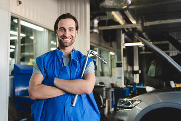 Smiling successful confident young male car technician mechanic in special blue robe uniform holding pipe monkey wrench looking at camera at vehicle service inspection.