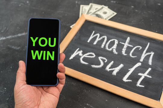 A hand with a smartphone on the screen with congratulations on the victory, a chalkboard with the text of the MATCH RESULT and dollar bills in the background.