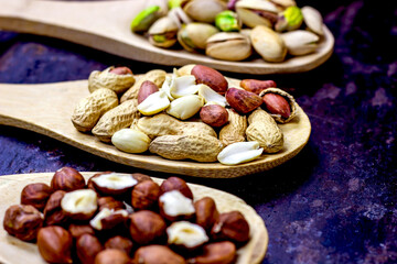 Different nuts: almonds, pistachios, peanuts, hazelnuts in light wooden spoons on black metal background.