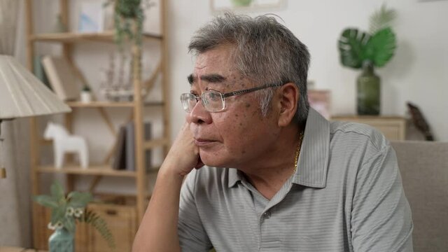 Senior asian man deep in thought while sitting on couch in living room at home. serious old male with hands putting under chin and feeling worried on sofa. sad people in glasses with upset emotion
