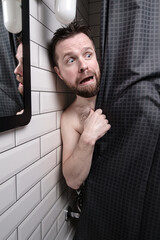 Wet, frightened man peeps out from behind the shower curtain and looks uneasy with a strange...