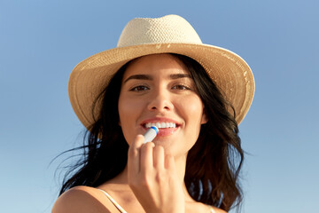 people, summer and swimwear concept - portrait of happy smiling young woman in bikini swimsuit and straw hat applying lip balm on beach - obrazy, fototapety, plakaty