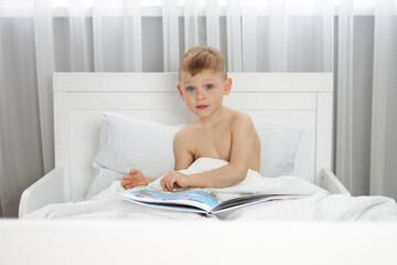 Fototapeta premium Little boy with blue eyes is reading a book in a white bed