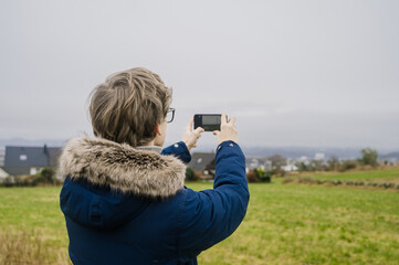 Obraz premium Young man in blue coat is taking a picture of city and green field by his phone. Cloudy day in Norway.