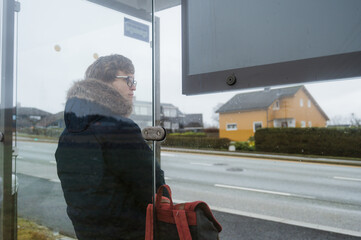 Man in blue coat with backpack is waiting for a bus. Modern glass bus stop on Norway. Cloudy day.