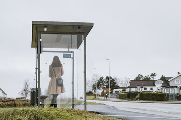 Elegant woman in coat is waiting for a bus. Modern glass bus stop on Norway. Cloudy day.