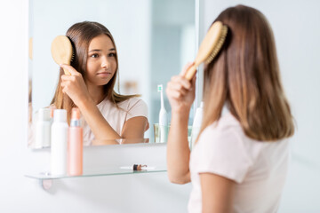 beauty and people concept - teenage girl looking to mirror and brushing hair with comb at home bathroom