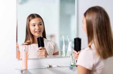 beauty and people concept - teenage girl looking to mirror and singing to hairbrush at bathroom