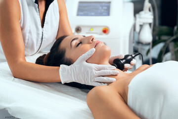 Cosmetologist makes the procedure an ultrasonic cleaning of the facial skin of a beautiful young woman on the spa center.