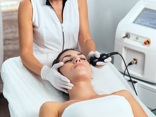 Cosmetologist makes the procedure an ultrasonic cleaning of the facial skin of a beautiful young woman on the spa center.
