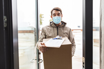 Fototapeta na wymiar health protection, safety and pandemic concept - delivery man in face protective mask holding parcel box with clipboard at open door