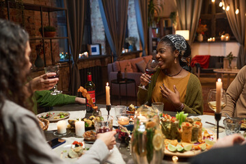 African young woman drinking red wine and laughing while having dinner with her friends at the table in the living room