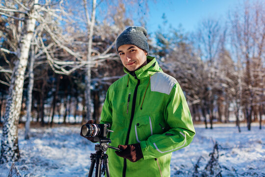 Young man photographer takes pictures of snowy winter forest using camera and tripod. Outdoor seasonal hobby activities
