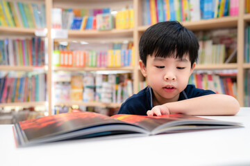 Adorable young Asia boy wearing blue shirt and sitting on the chair in the modern library or bookstore and reading a book. Education and learning concept. Kid interest cartoon book. Feeling happy