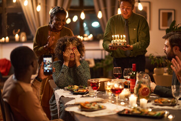 Group of friends congratulating woman with her birthday, they carrying birthday cake with candles while sitting at the table with her eyes closed